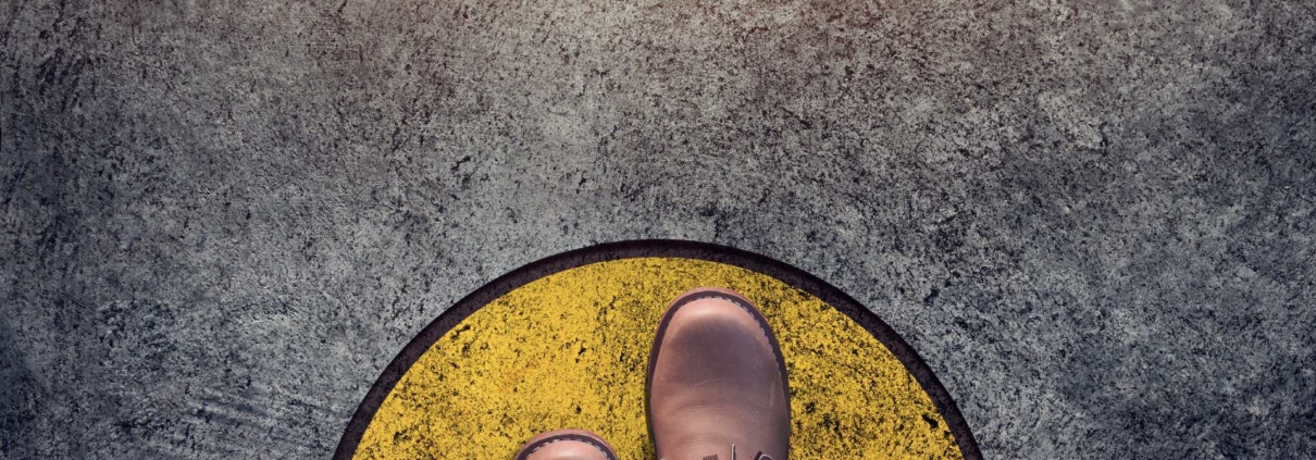 A view from someone looking down at their shoes: manly brown dress shoes stepping on a floor that has a yellow circle with a black outline painted on the concrete grey floor