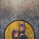 A view from someone looking down at their shoes: manly brown dress shoes stepping on a floor that has a yellow circle with a black outline painted on the concrete grey floor